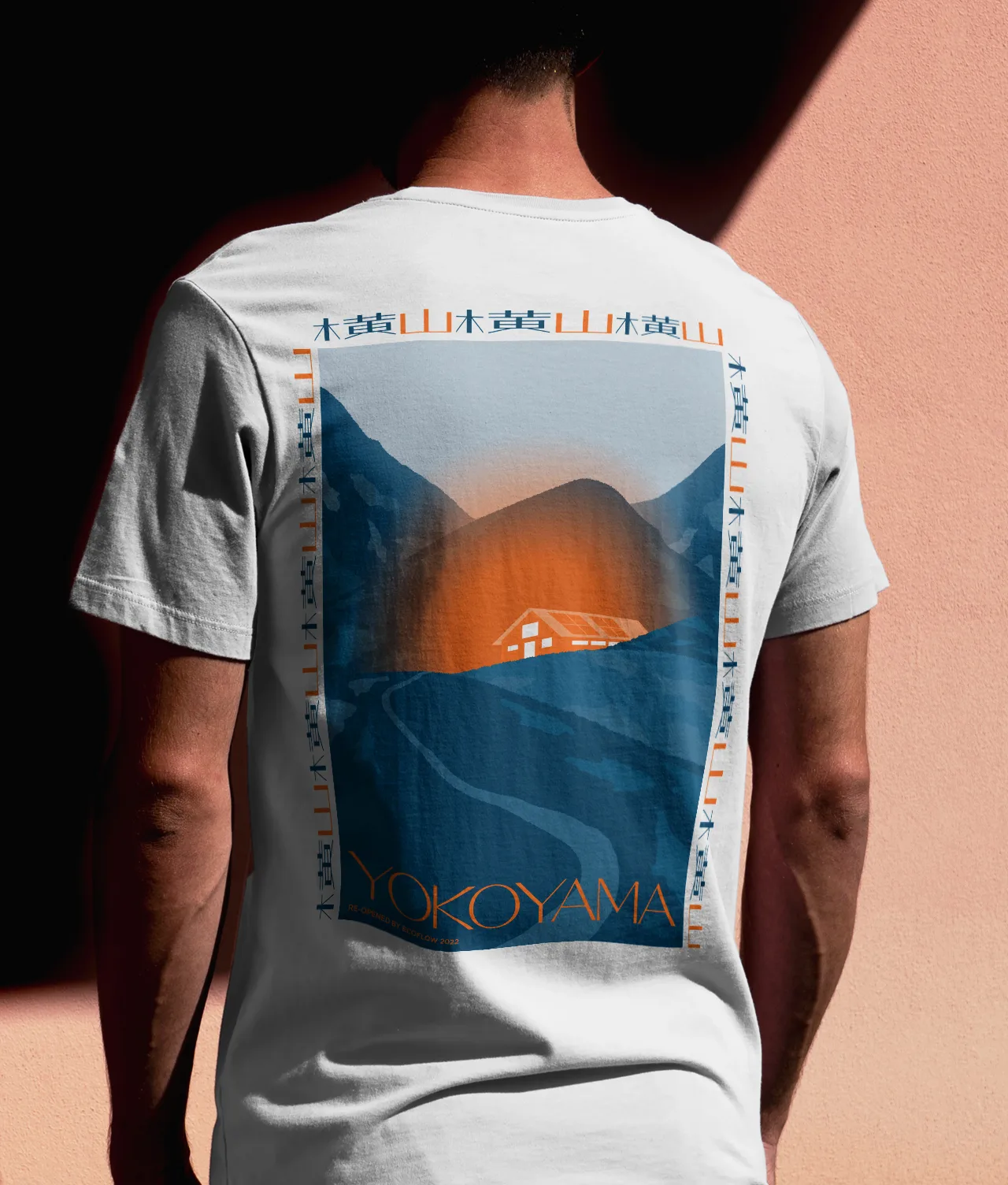 A person wearing a white T-shirt with a large, stylized graphic on the back depicting mountains, a sunset, and a house, with the word YOKOYAMA at the bottom. The person stands against a peach-colored wall.