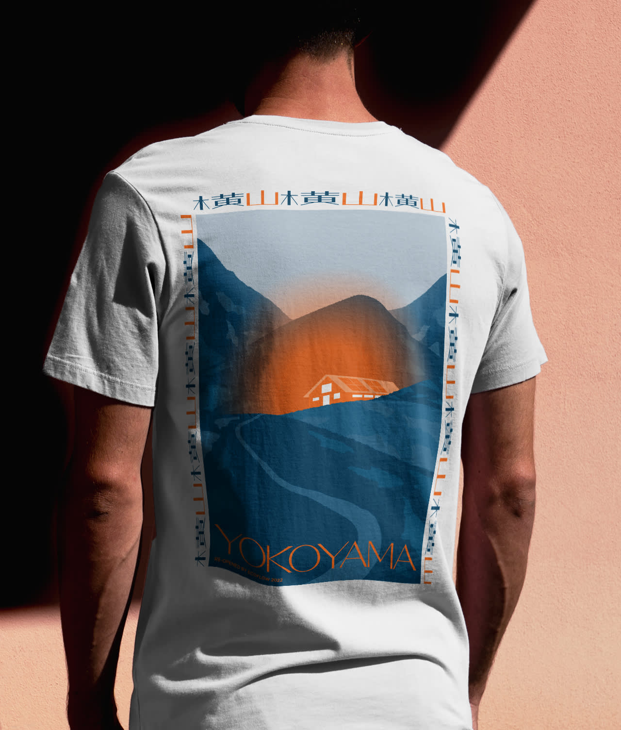 A person wearing a white T-shirt with a large, stylized graphic on the back depicting mountains, a sunset, and a house, with the word YOKOYAMA at the bottom. The person stands against a peach-colored wall.