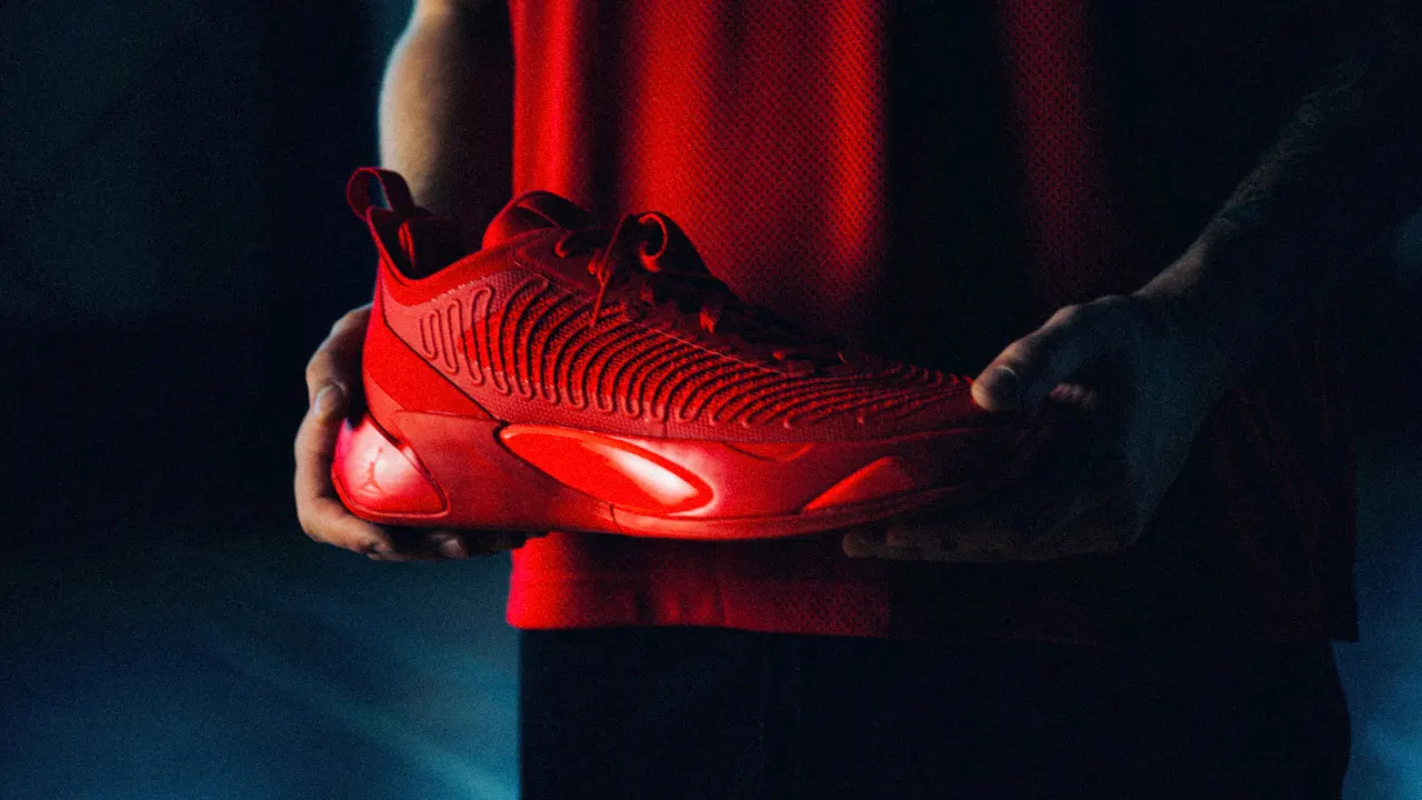 A person in a red shirt holds a bright red athletic sneaker with textured details, illuminated against a dark, blurred background.