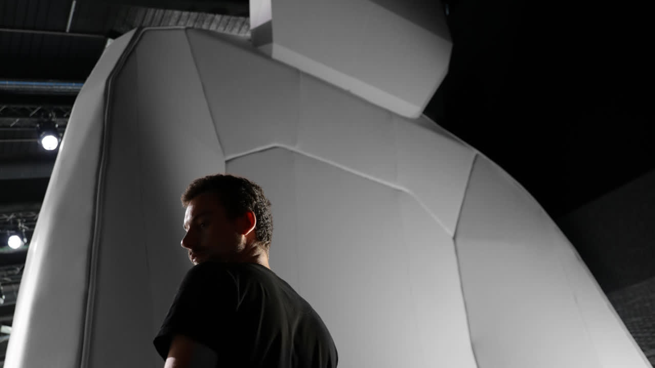 A man in a black shirt stands in front of a large, abstract, geometric sculpture resembling a seated figure, with dramatic lighting and shadows emphasizing the scale and shape.