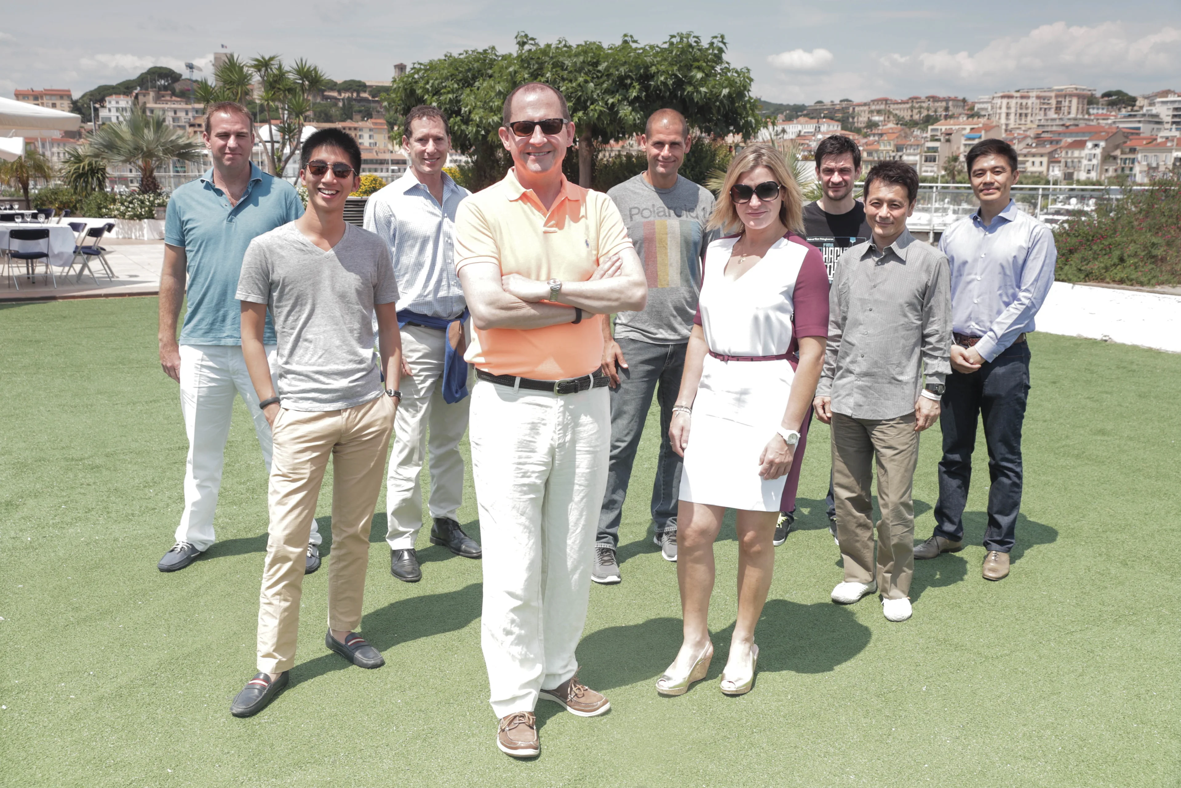 A group of ten people, eight men and two women, stand outside on grass with buildings and palm trees in the background. The individuals are smiling and dressed in casual and business-casual attire.