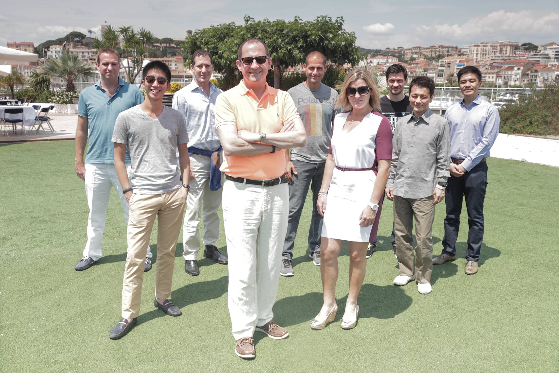 A group of ten people, eight men and two women, stand outside on grass with buildings and palm trees in the background. The individuals are smiling and dressed in casual and business-casual attire.
