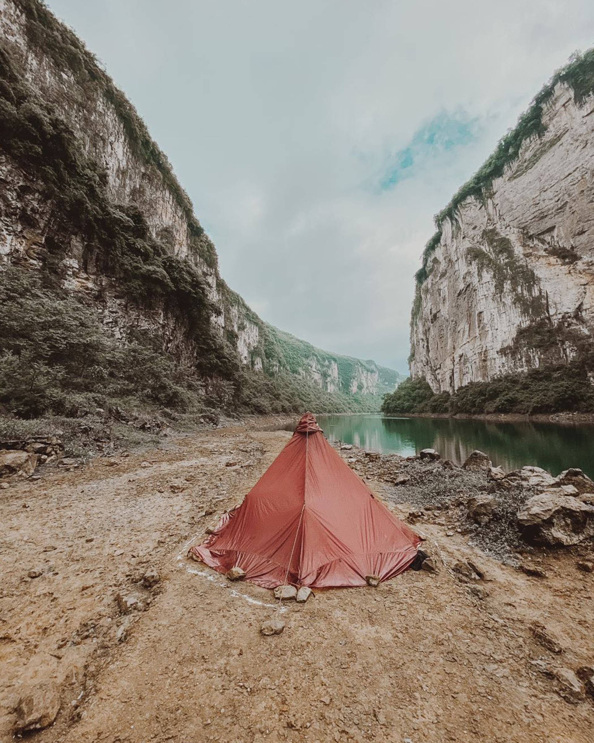 A red tent is set up on rocky ground beside a calm river, surrounded by steep, green cliffs under a partly cloudy sky.