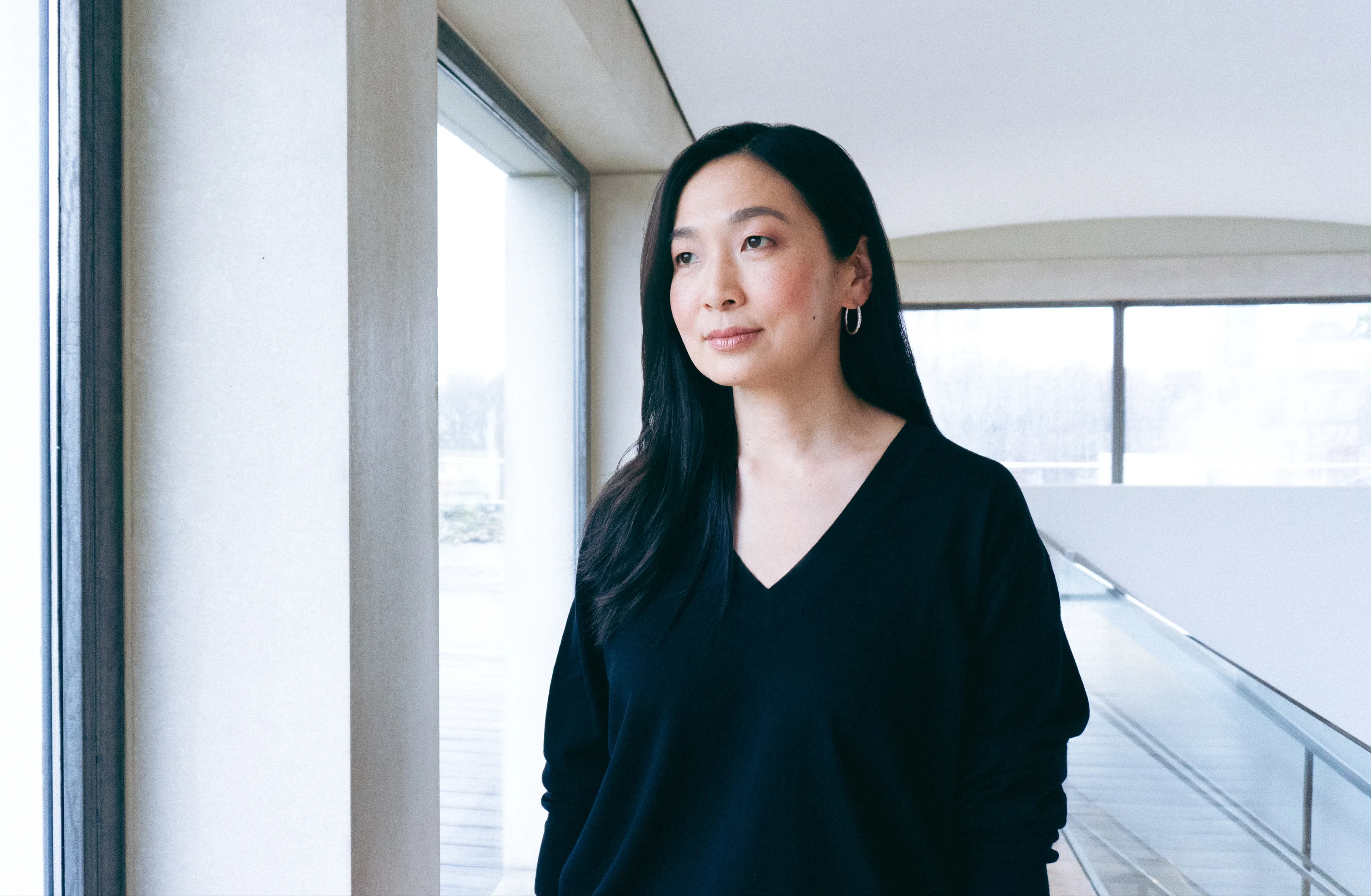 A woman with long dark hair wearing a black V-neck top stands indoors by large windows, looking outside thoughtfully. The background shows a modern, bright interior space with natural light.