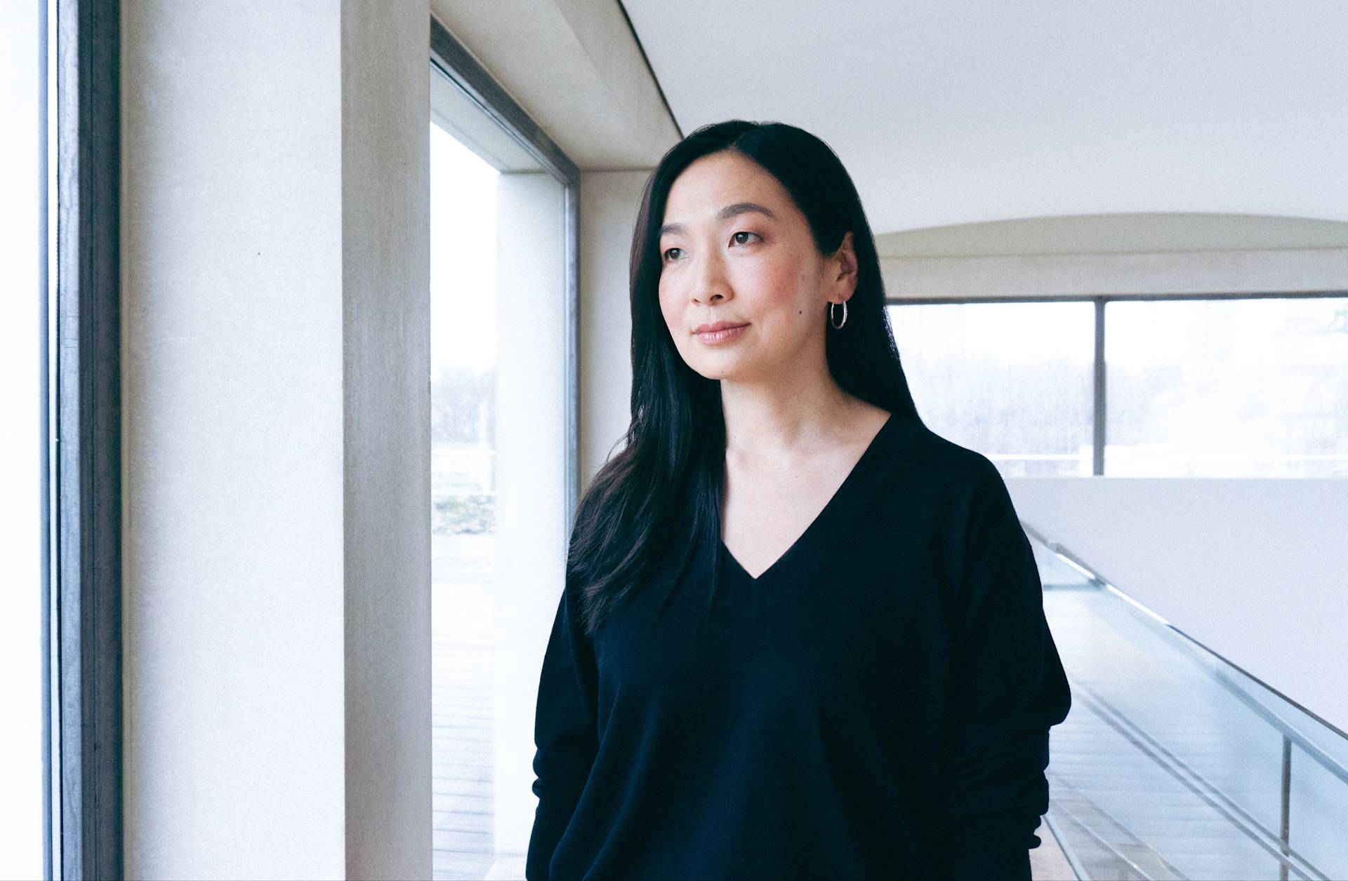 A woman with long dark hair wearing a black V-neck top stands indoors by large windows, looking outside thoughtfully. The background shows a modern, bright interior space with natural light.