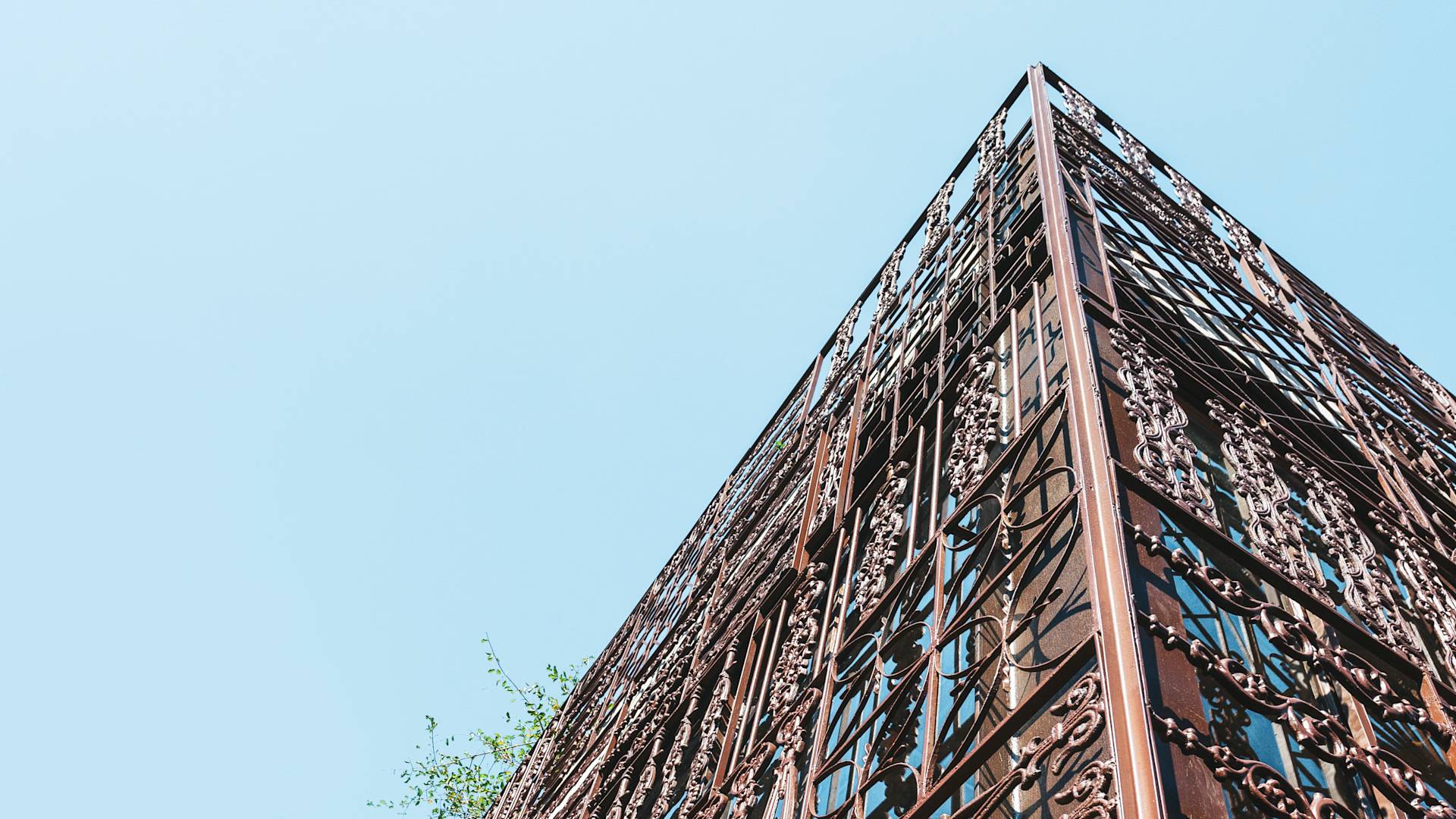 A low-angle view of a building’s corner with intricate, decorative metalwork on its facade, set against a clear, pale blue sky. Some greenery is slightly visible on the left side.