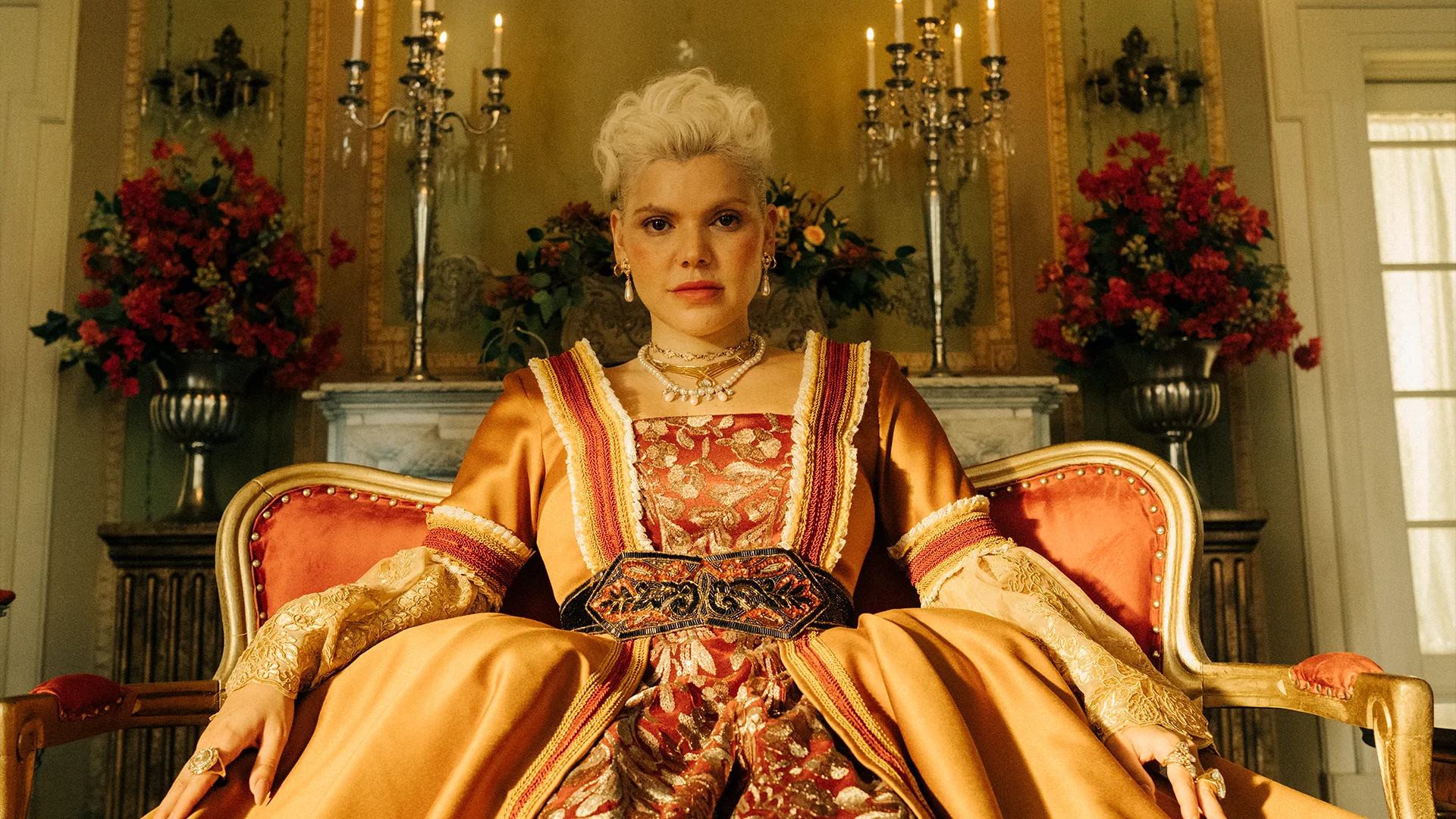 A person with short, styled white hair wears an ornate gold and red gown, sitting confidently on a vintage armchair in an elegant room with chandeliers, flowers, and gold decor.