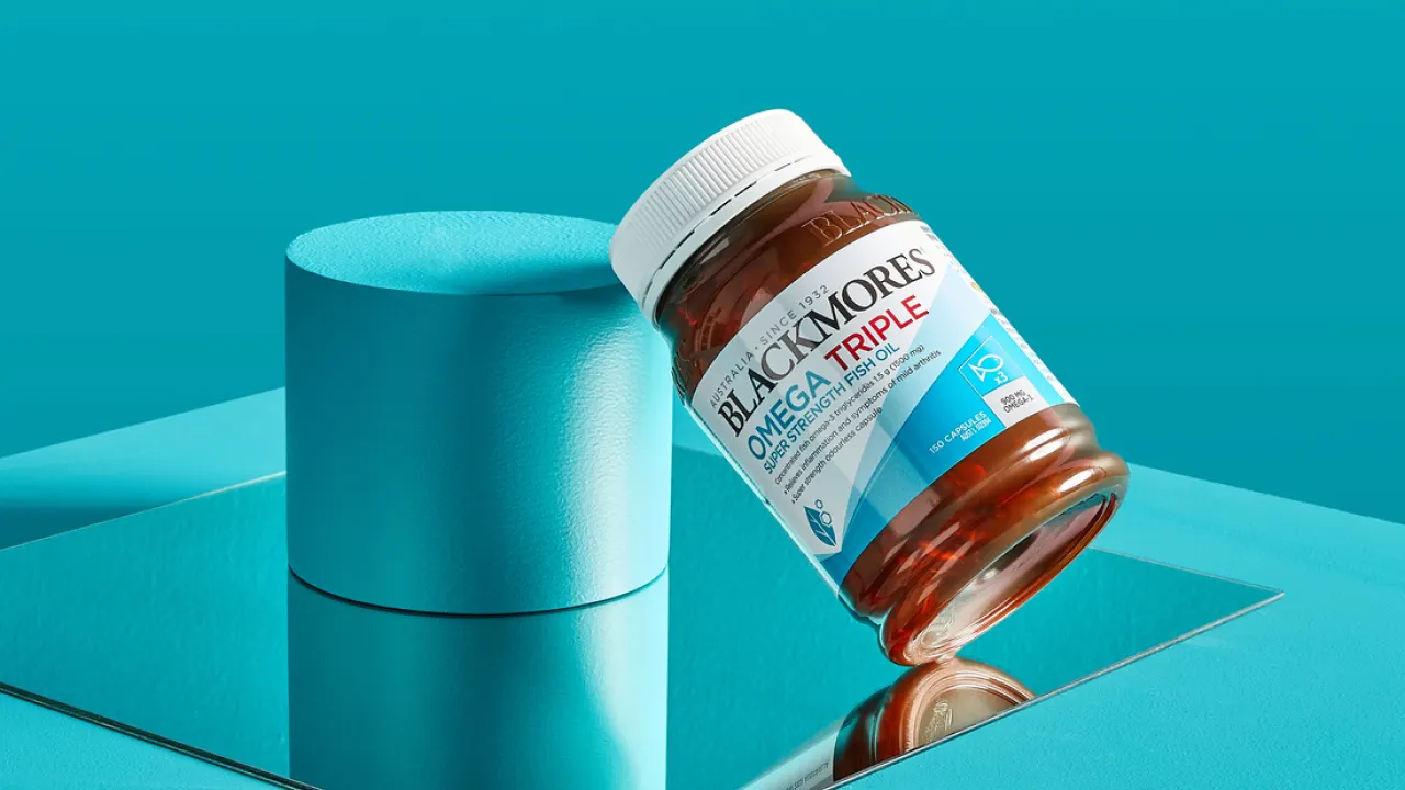 A bottle of Blackmores Omega Triple Fish Oil capsules rests at an angle on a blue cylindrical pedestal, with a blue background and a mirror reflecting the bottle and pedestal.
