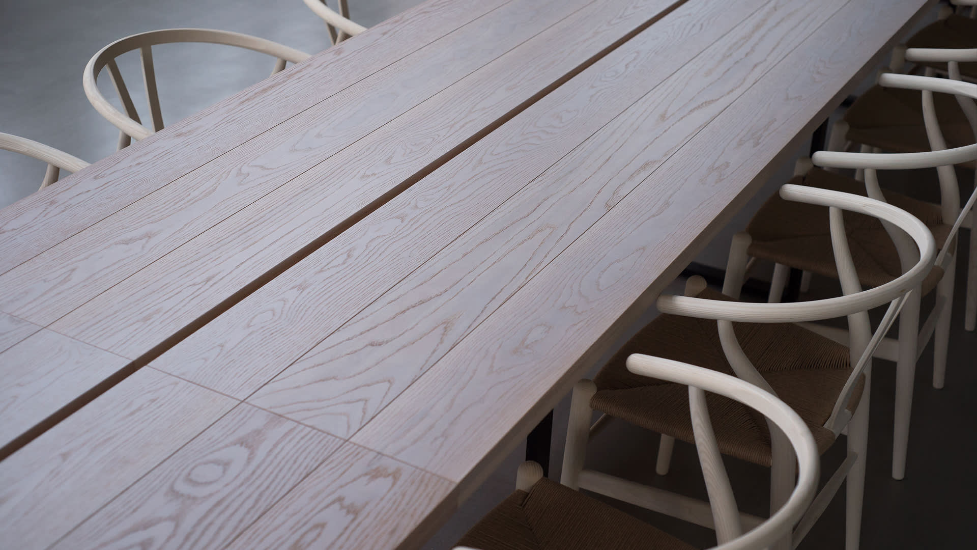 A long, light-colored wooden table with visible grain patterns is surrounded by several modern, light chairs featuring curved backs and woven seats. The setting appears minimalist and uncluttered.