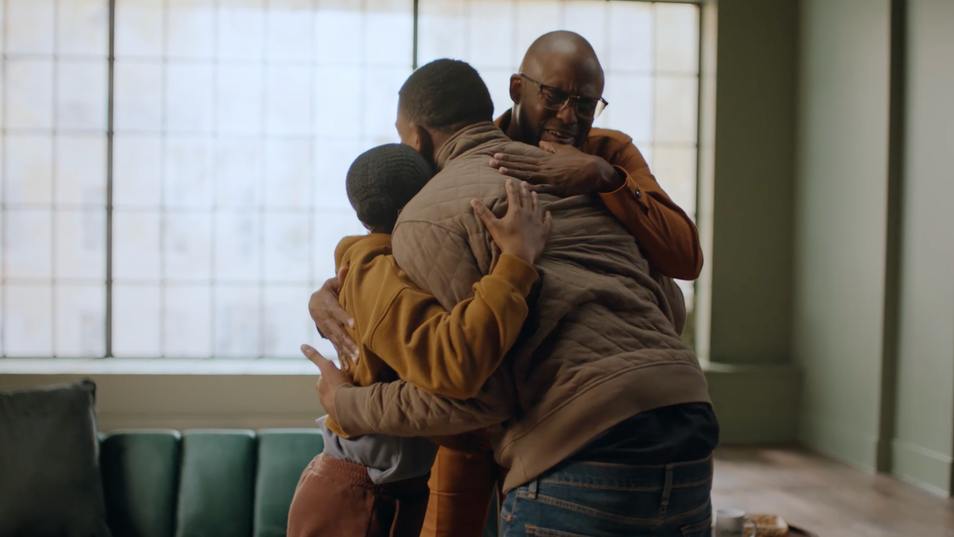 Three people stand indoors, embracing each other warmly in a group hug. One person has their eyes closed, smiling, while sunlight filters through large windows in the background.