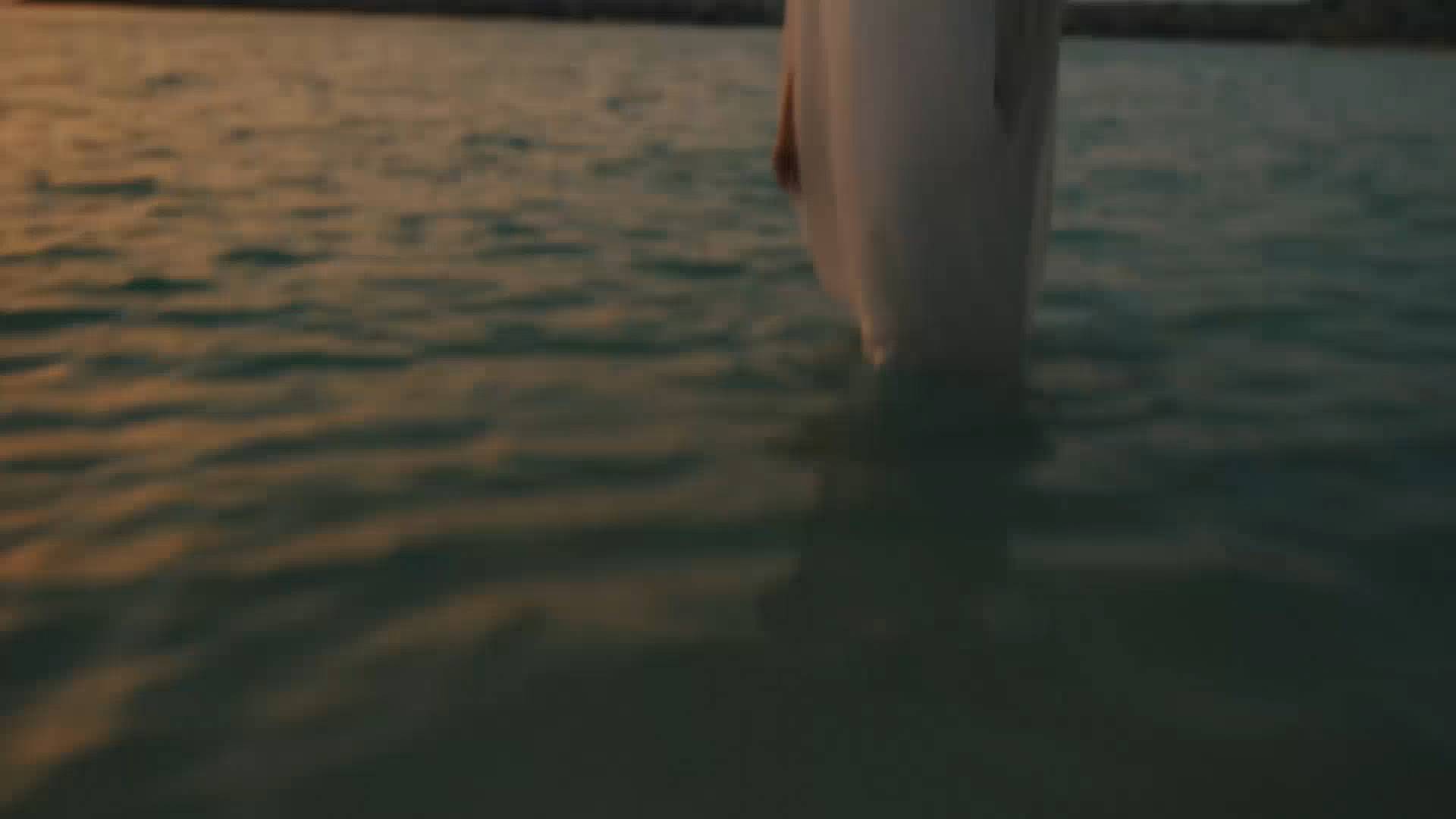 A person in a flowing white dress stands in shallow water at sunset, with gentle ripples around their legs and warm light reflecting off the surface.