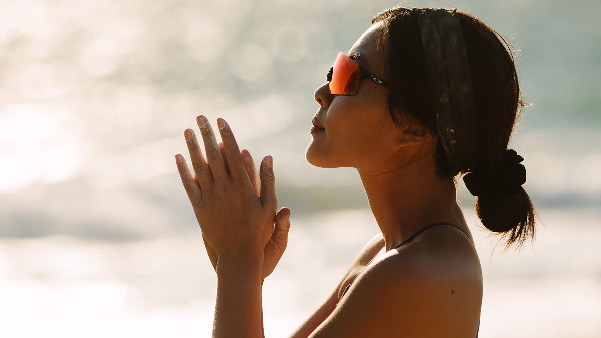 A woman wearing sunglasses and a headband stands sideways by the beach, hands pressed together in a prayer or meditation pose, with sunlight reflecting off the water in the background.