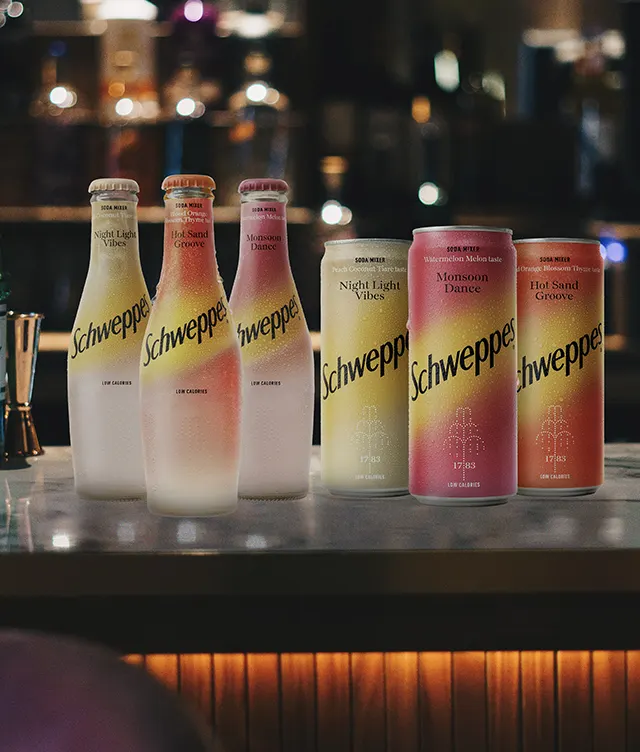 Four glass bottles and four cans of Schweppes sodas in pink and yellow gradients are arranged on a bar counter, illuminated by warm lighting with a blurred background of bottles and bar decor.