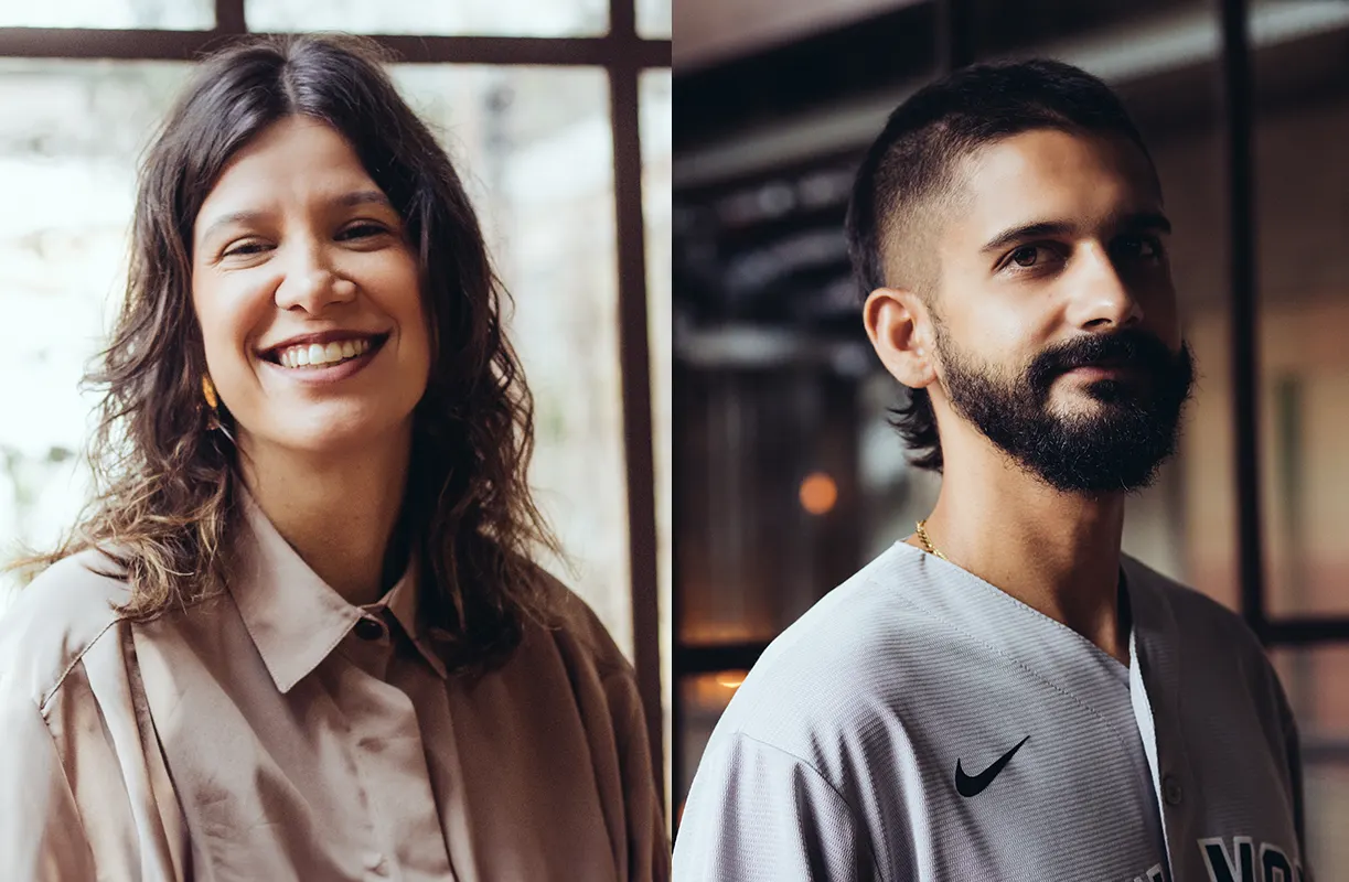 A woman with wavy hair smiles, wearing a beige shirt. Next to her, a man with short dark hair, a beard, and a mustache looks at the camera, wearing a light gray Nike shirt. Both are indoors with blurred backgrounds.