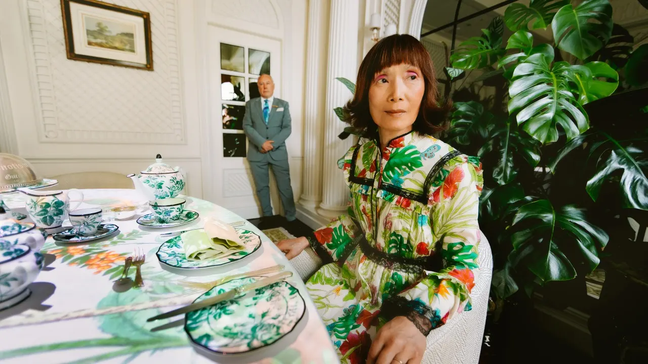 A woman in a colorful floral dress sits at an elegantly set table with green and white china. A man in a suit stands in the background, and large leafy plants decorate the bright, ornate room.