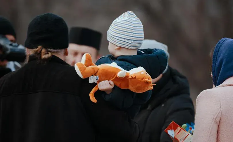 A child wearing a striped beanie and dark jacket holds an orange stuffed animal while standing among adults dressed in winter clothing. The background is blurred.