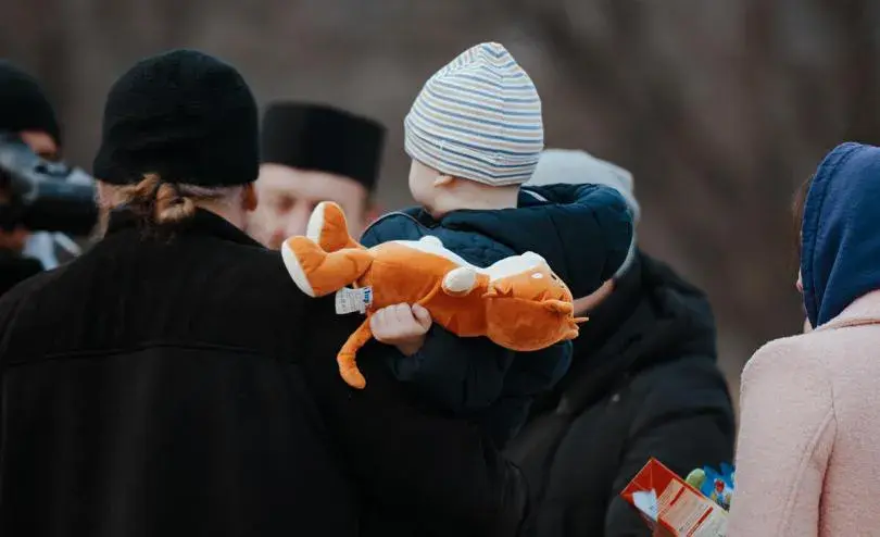A child wearing a striped beanie and dark jacket holds an orange stuffed animal while standing among adults dressed in winter clothing. The background is blurred.