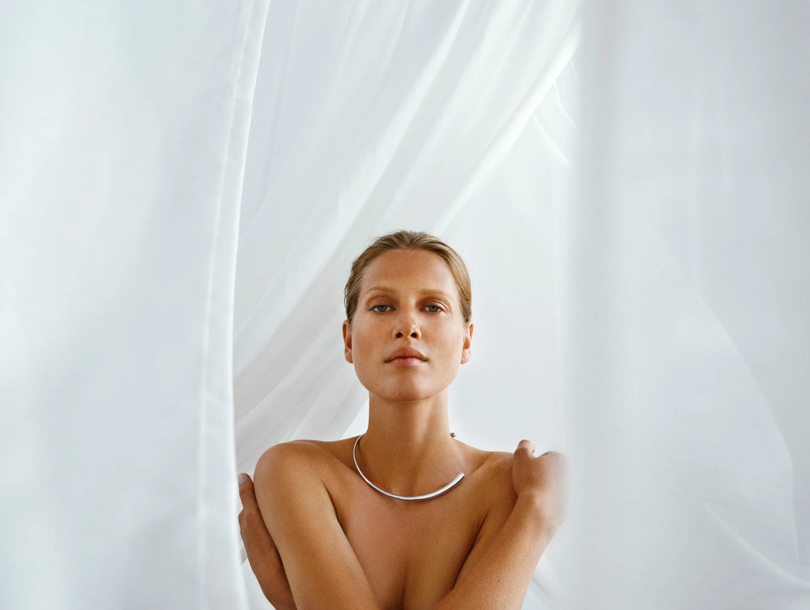 A woman with bare shoulders and a silver necklace stands in front of sheer white curtains, softly lit by natural light, looking directly at the camera with a calm expression.