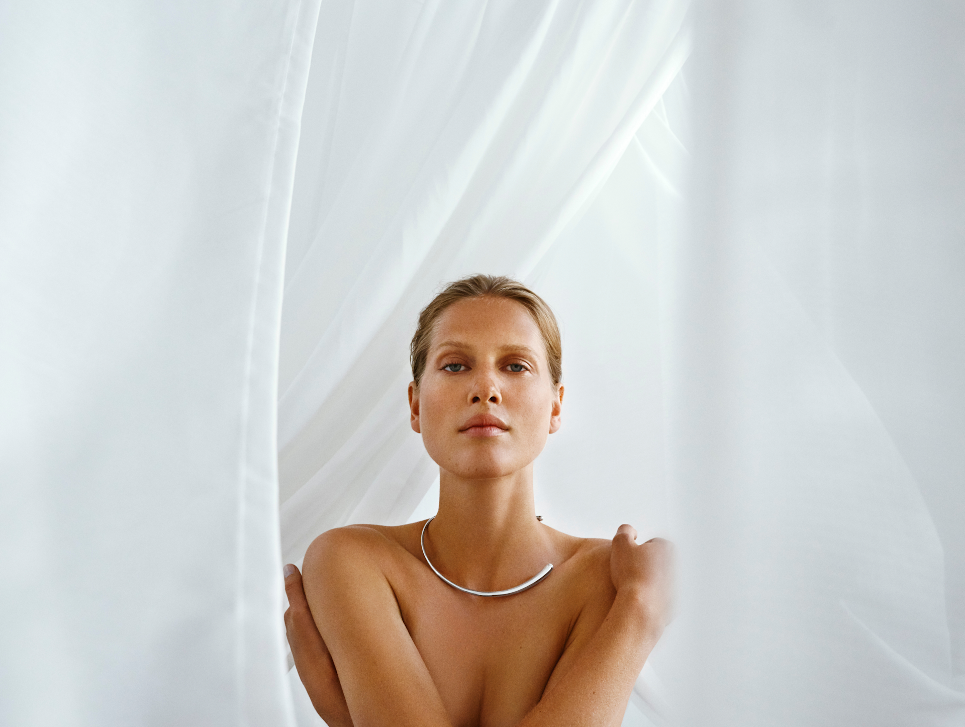 A woman with bare shoulders and a silver necklace stands in front of sheer white curtains, softly lit by natural light, looking directly at the camera with a calm expression.