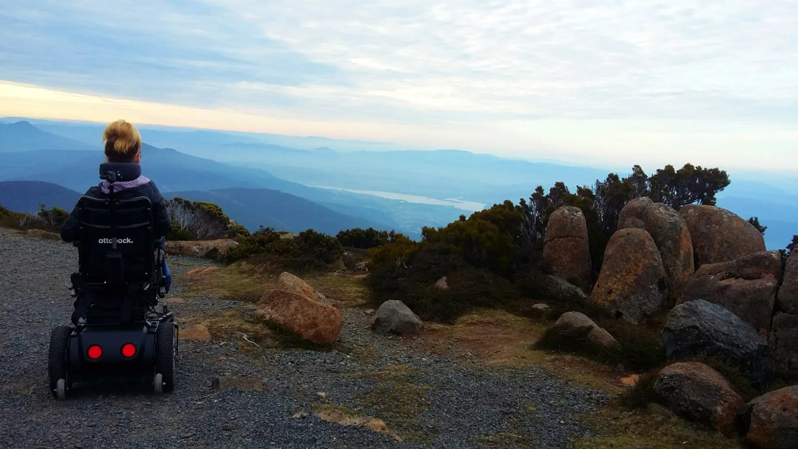 A person in a powered wheelchair looks out over a scenic mountain landscape with rolling hills, rocky terrain, and a cloudy sky.