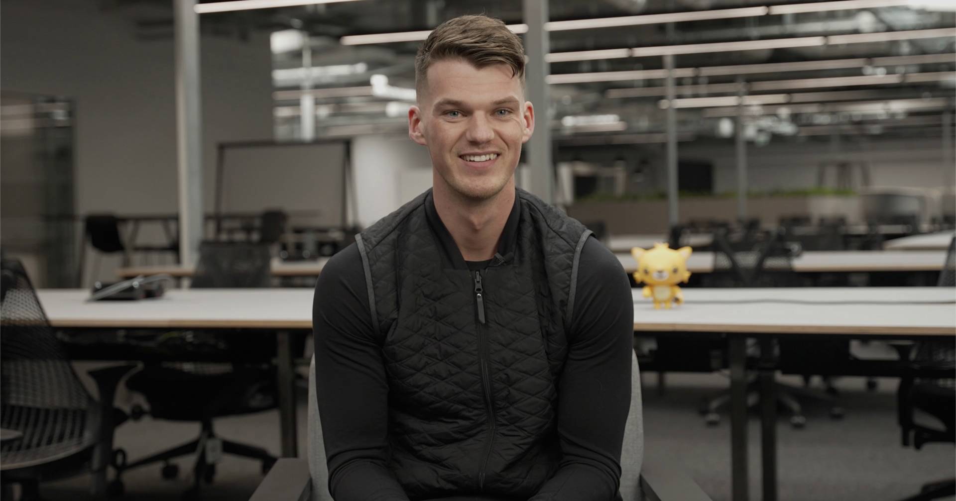 A man with short brown hair wearing a black quilted vest and long-sleeve shirt sits and smiles in a modern office with empty desks. A yellow toy figure is on the table behind him.