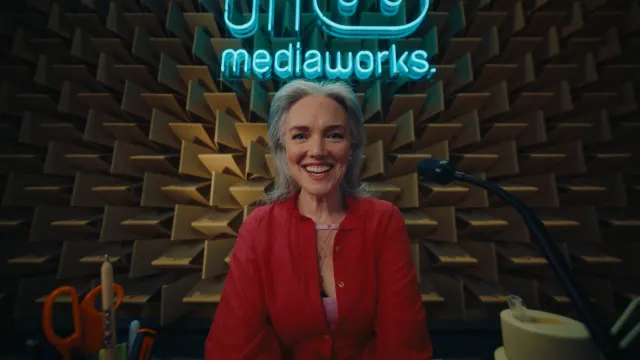 A smiling woman with gray hair and a red shirt sits in front of a microphone in a soundproof studio, with a neon “mediaworks” sign and acoustic foam panels behind her. Office supplies are on the desk in front.
