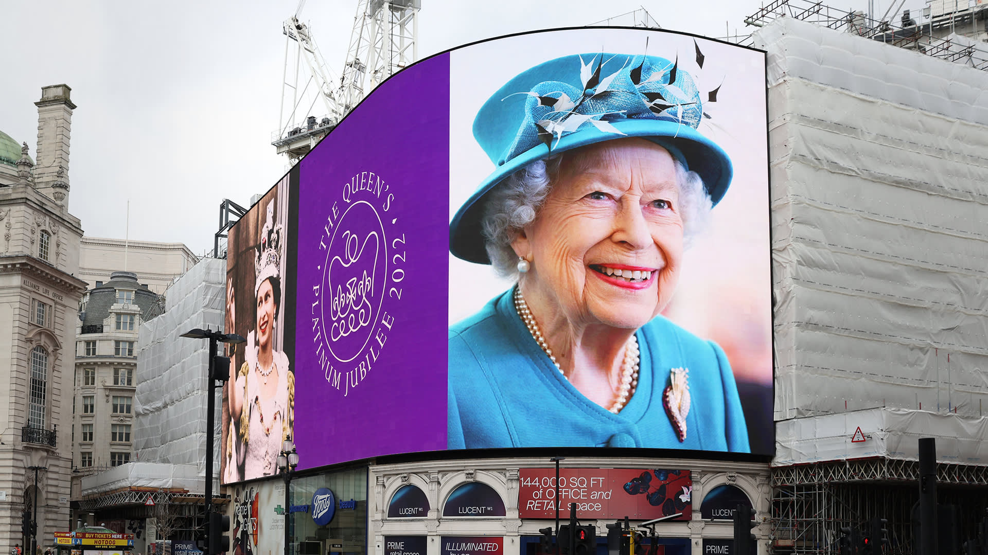 A large digital billboard displays a portrait of Queen Elizabeth II and a purple emblem reading The Queen’s Platinum Jubilee 2022 in a busy city square with nearby construction and historic buildings.