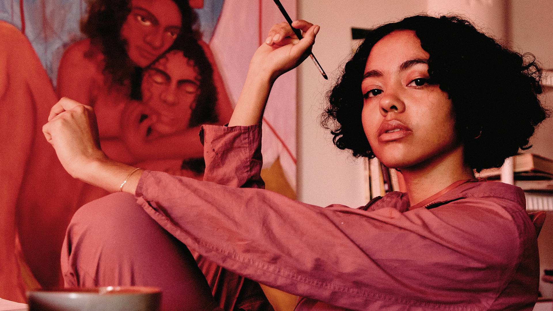 A young person with curly hair and a serious expression sits indoors, holding a paintbrush. They wear a pinkish outfit, and behind them is a colorful painting featuring two figures. A mug is visible in the foreground.