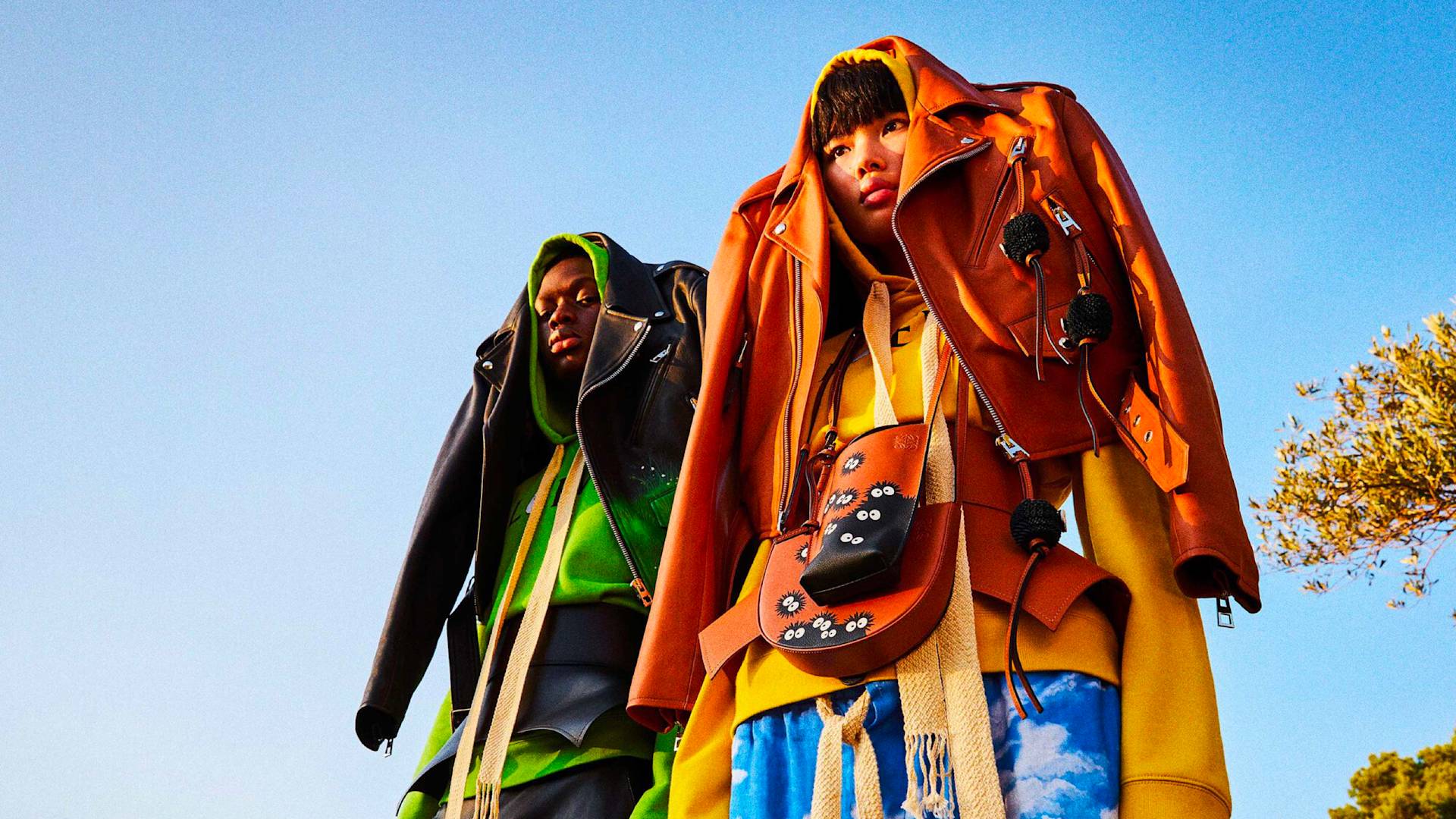 Two people stand outdoors under a blue sky, each wearing layered, oversized jackets draped over their heads and colorful outfits, with creative accessories and a tree partially visible to the right.