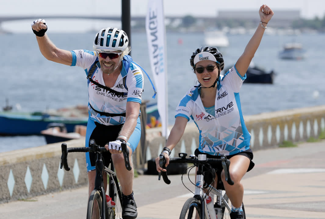 Two cyclists, a man and a woman, wearing matching blue and white jerseys and helmets, ride side by side along a waterfront path, smiling and raising their arms in celebration. Boats and water are visible in the background.
