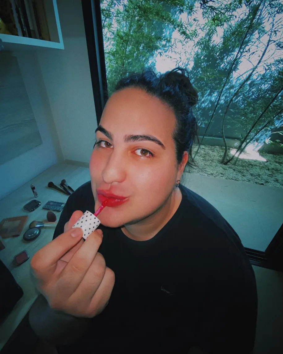 A person with dark hair in a bun is applying red lipstick while sitting indoors near a large window with trees visible outside. Makeup items are scattered on the table beside them.