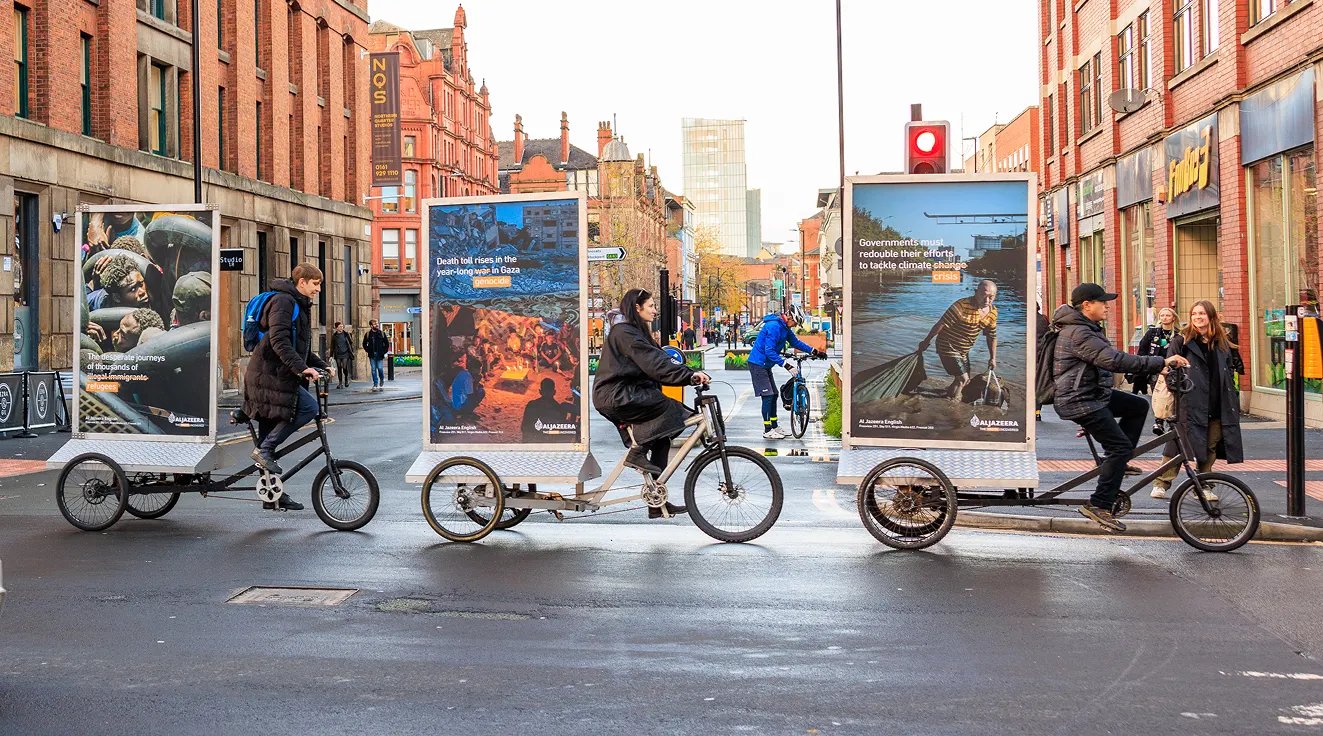 People ride custom tall bicycles across a city street, passing by large posters with animal and nature images. Two women stand on the sidewalk, and buildings line both sides of the street under an overcast sky.