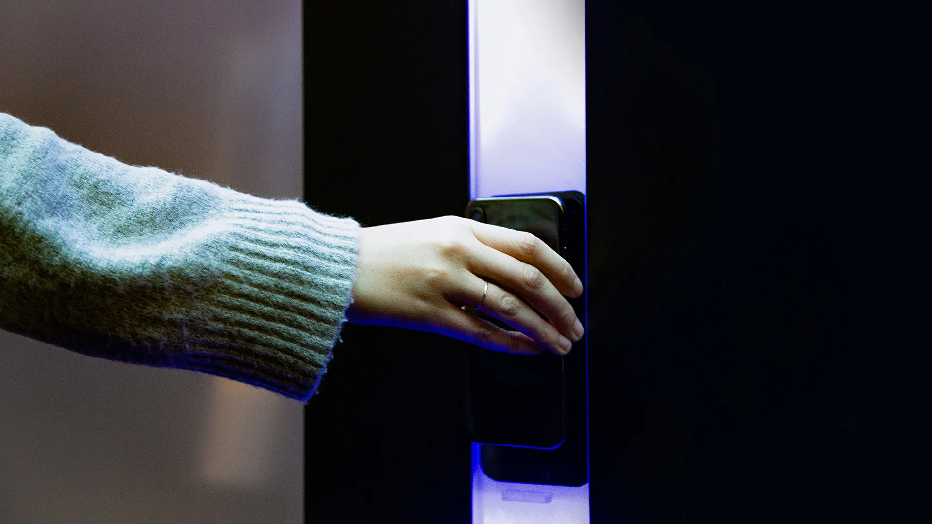 A person’s hand in a gray sweater holding a smartphone against a card reader or access panel, illuminated by blue light, suggesting contactless entry or digital authentication.