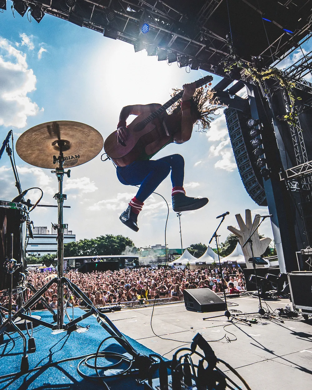 A musician jumps mid-air while playing guitar on an outdoor stage, with a drum set nearby and a large crowd cheering under a sunny sky.