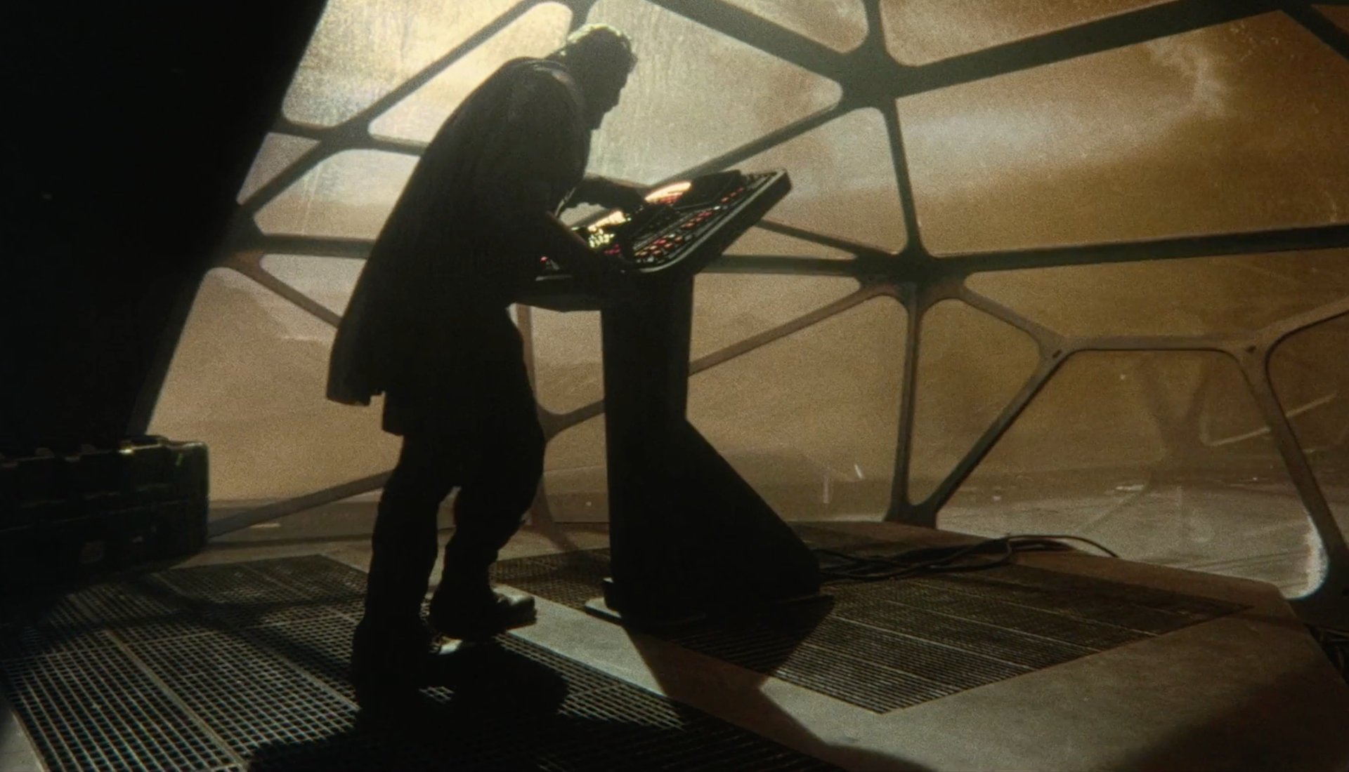A person in shadow operates a futuristic control panel inside a dimly lit, industrial room with hexagonal window patterns and a dusty, golden atmosphere outside.