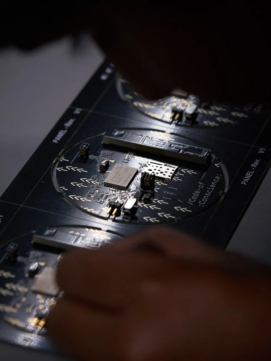 Close-up of a person examining an electronic circuit board with various components, illuminated under focused lighting, with another similar board visible in the background.