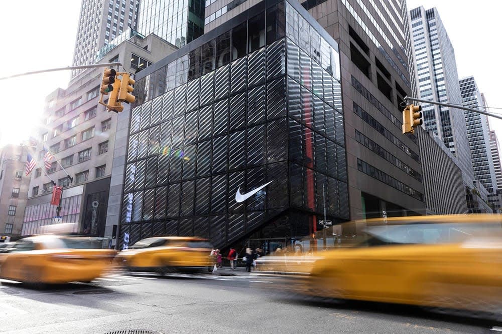 A Nike store with a large white swoosh logo on a glass building corner in a busy city, with yellow taxis and blurred traffic passing by on the street. Tall skyscrapers surround the area.