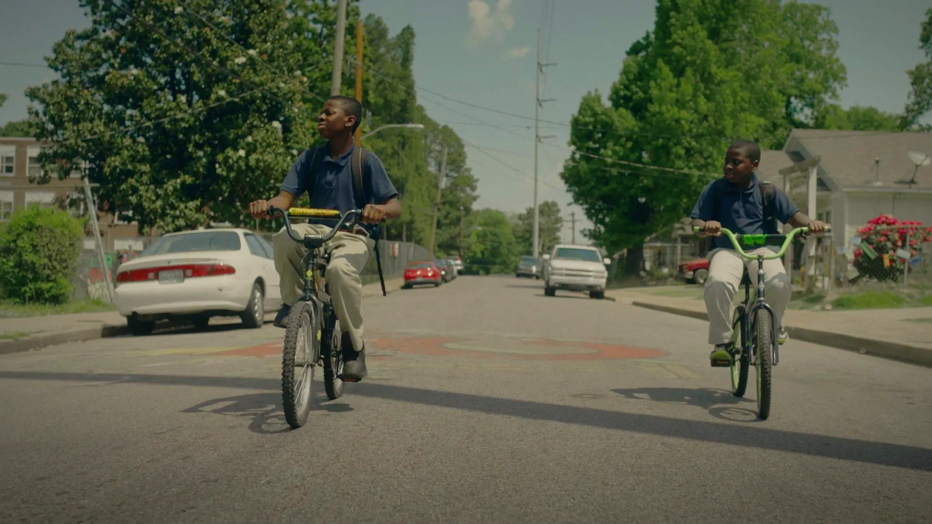 Two boys ride bicycles down a quiet residential street on a sunny day. They wear dark blue shirts and khaki pants, and houses, cars, and green trees line both sides of the road.