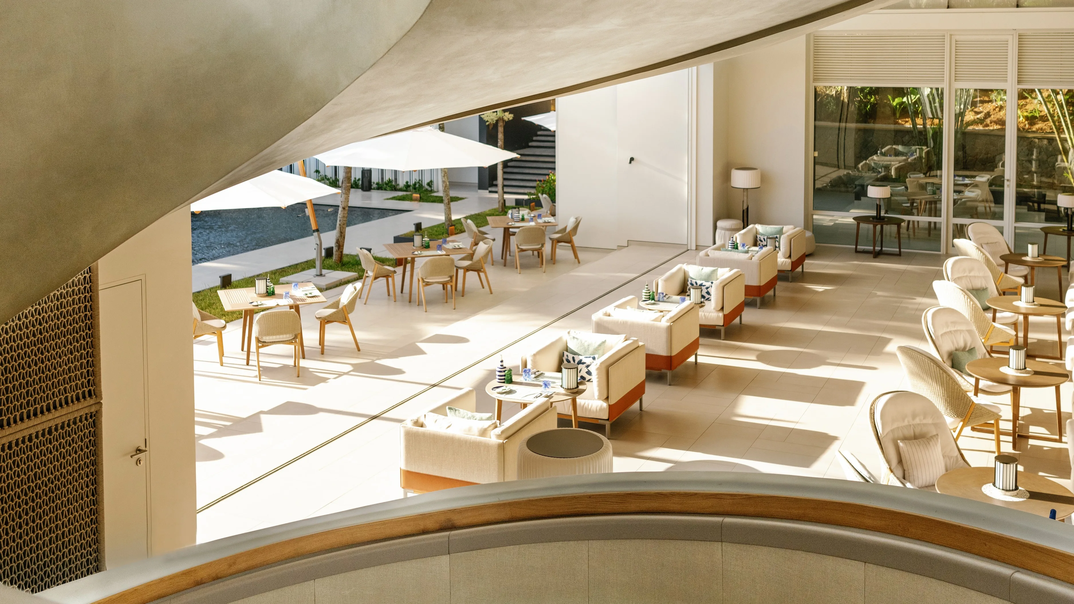 Modern, sunlit indoor-outdoor dining area with cream-colored chairs and tables, open to a patio with umbrellas and poolside seating, seen from a curved staircase. The space looks elegant, airy, and inviting.