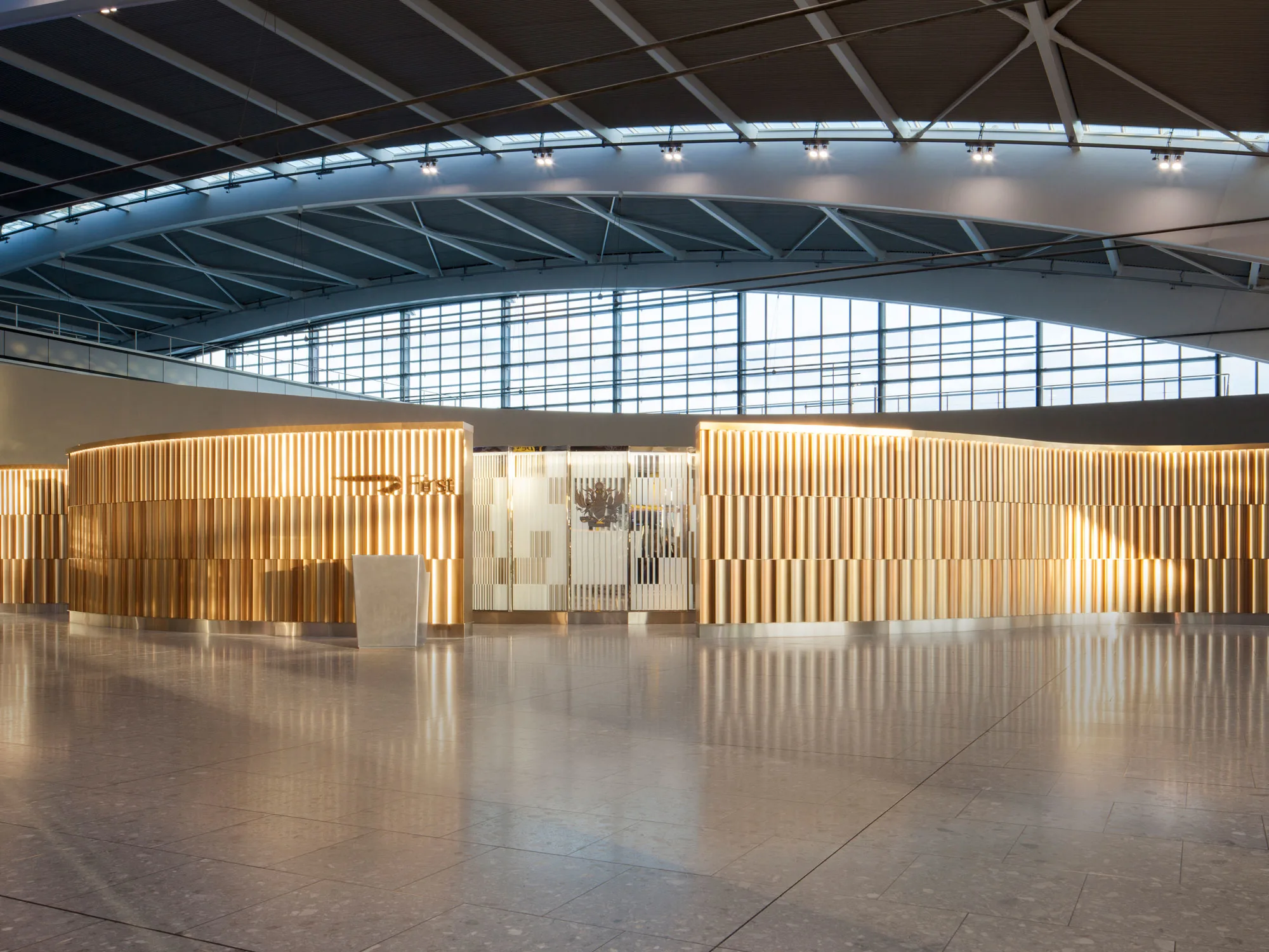 A modern airport lounge entrance with a curved, gold-toned facade, a minimalist white reception desk, and large windows letting in natural light under an arched ceiling.