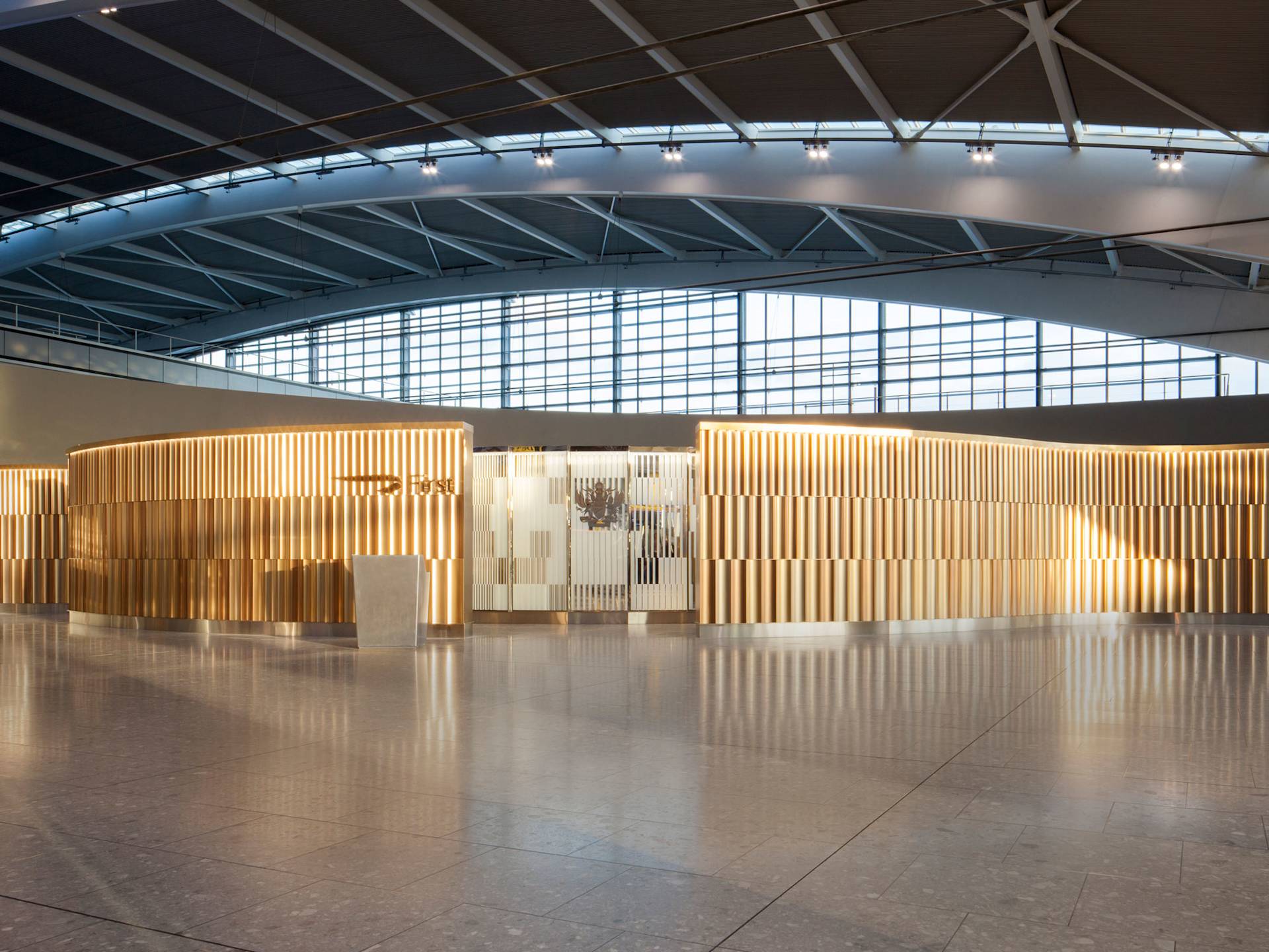 A modern airport lounge entrance with a curved, gold-toned facade, a minimalist white reception desk, and large windows letting in natural light under an arched ceiling.