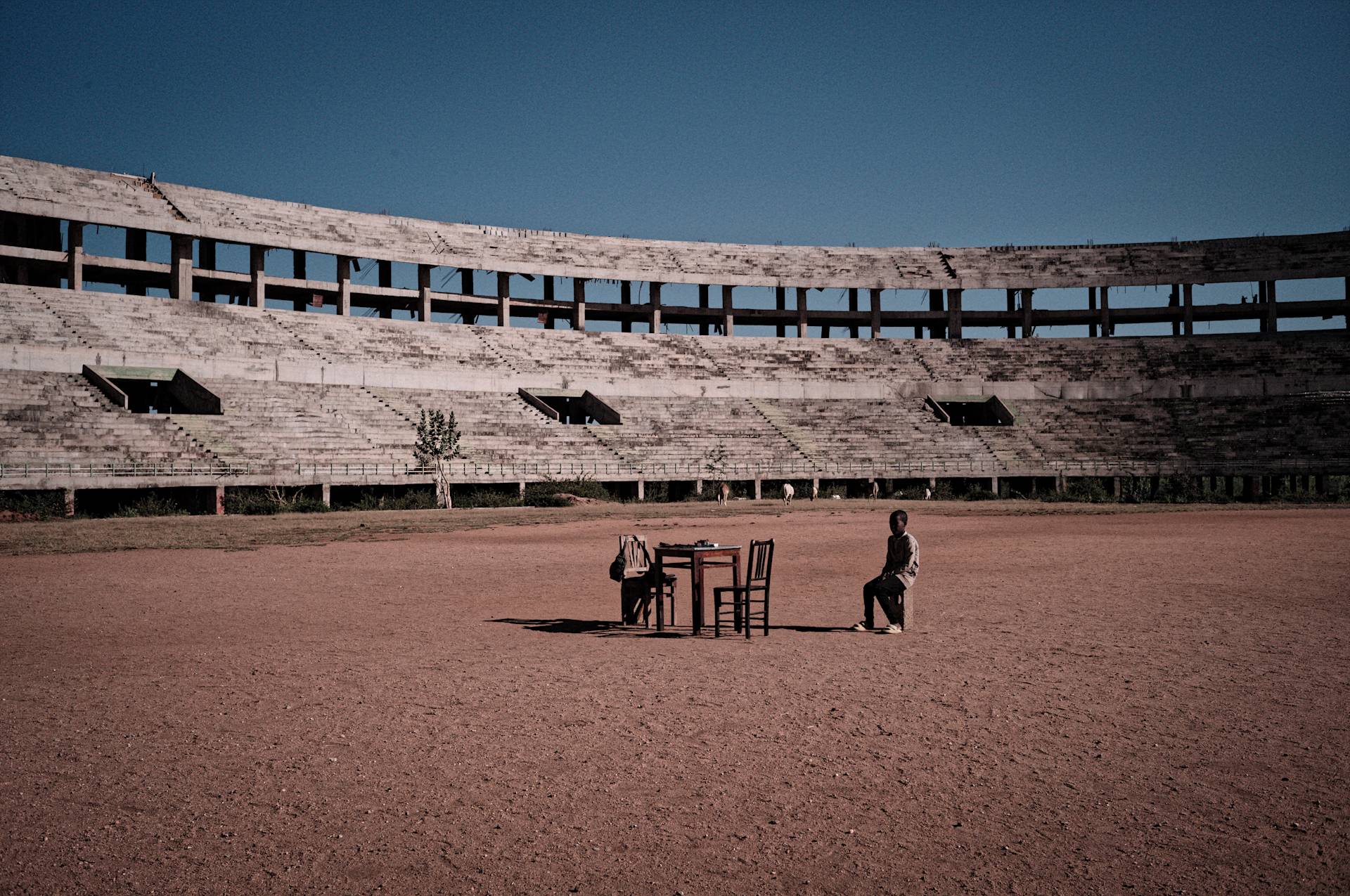 A person sits alone at a table and chair set in the center of an empty, weathered stadium under a clear blue sky. The stadium seating is old and partially overgrown with weeds.