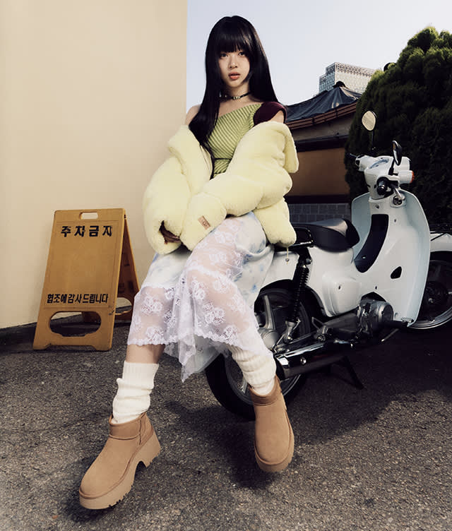 A woman with long black hair sits on a white scooter. She wears a yellow puffer jacket, green top, sheer white lace skirt, white leg warmers, and tan boots. A yellow caution sign with Korean text stands nearby.