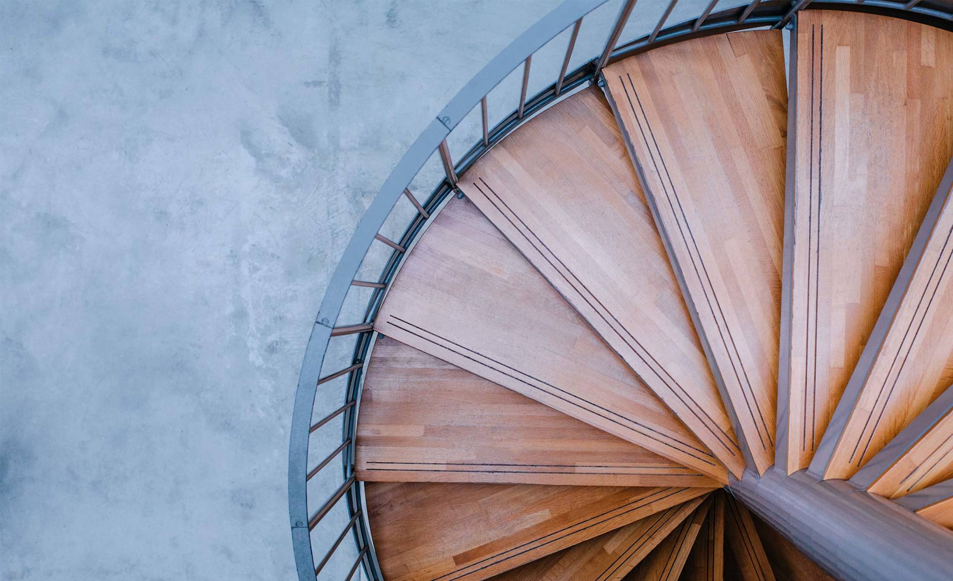 A top-down view of a wooden spiral staircase with metal railings, set against a smooth, light gray concrete floor. The stairs curve around a central pole, creating a geometric, fan-like pattern.