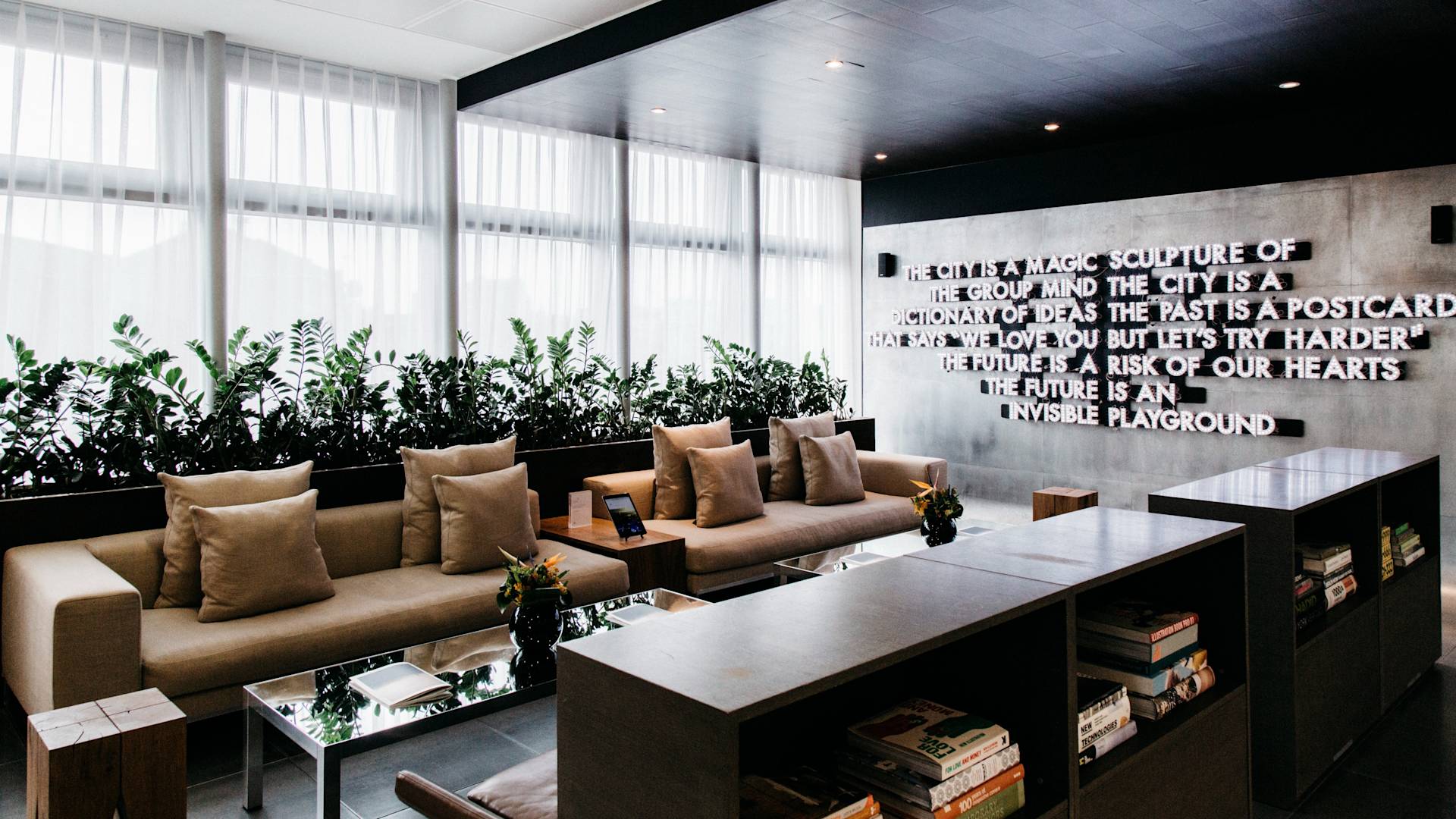 Modern lounge with beige sofas, glass tables, potted plants, and bookshelves. Large windows with sheer curtains let in natural light. A wall features a bold, artistic text installation. Decorative flowers are on the tables.