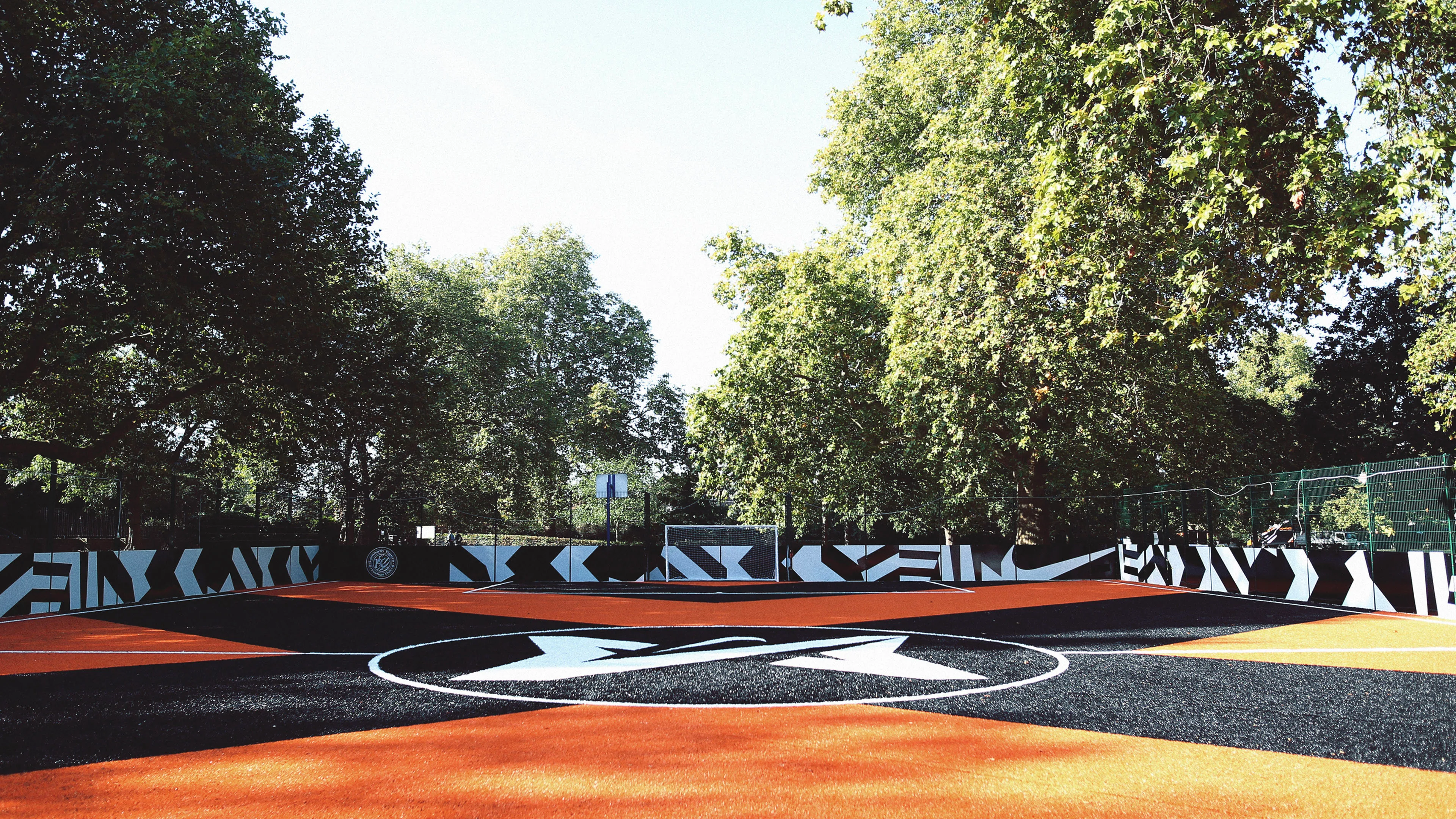 A vibrant outdoor soccer field with a black and orange patterned surface and bold geometric designs along the surrounding fences, set among tall green trees under a clear sky.