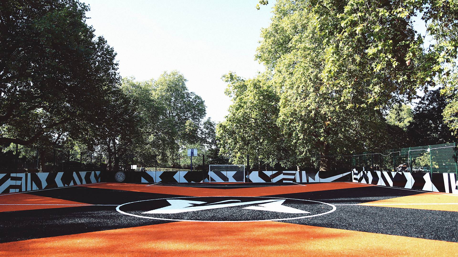 A vibrant outdoor soccer field with a black and orange patterned surface and bold geometric designs along the surrounding fences, set among tall green trees under a clear sky.