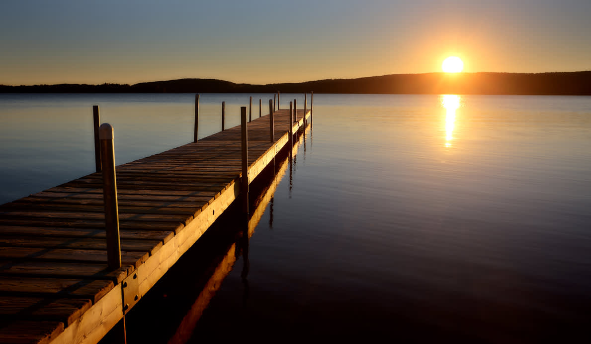 A wooden dock extends into a calm lake at sunset, with the sun low over distant hills and its reflection shimmering on the water.