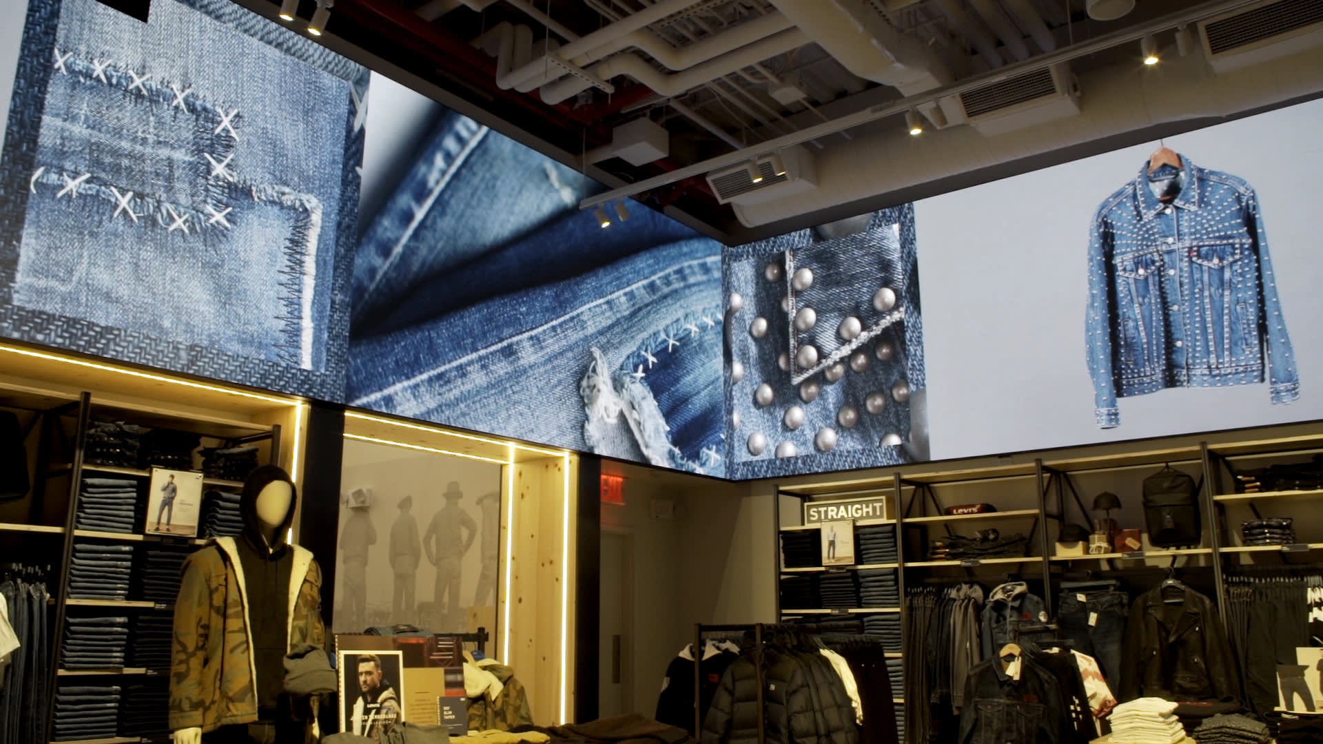 A clothing store displays denim jeans and jackets on shelves and mannequins, with a large digital screen above showing close-ups and a studded denim jacket. The store has bright lighting and visible ceiling pipes.