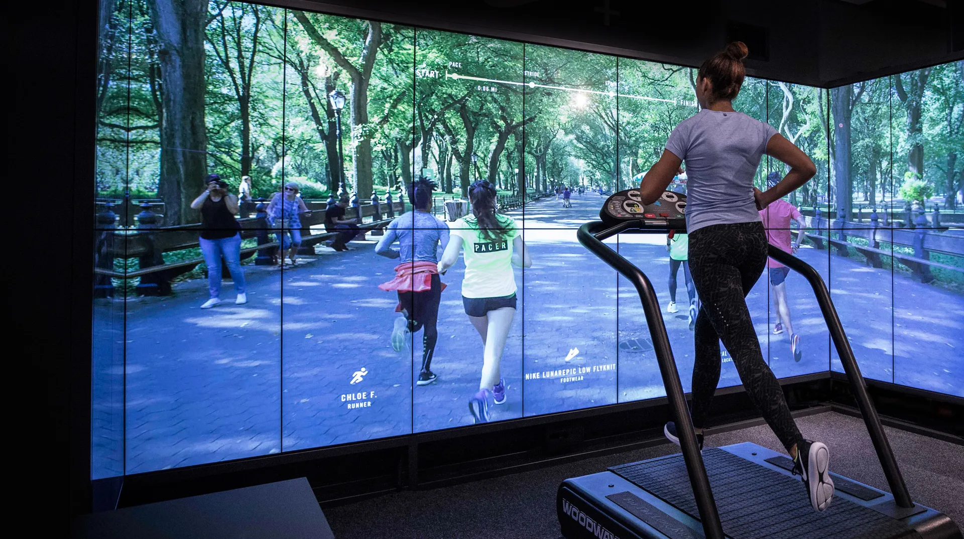 A woman runs on a treadmill facing large screens displaying a virtual outdoor park scene with people jogging, creating the illusion of running outdoors inside a gym.